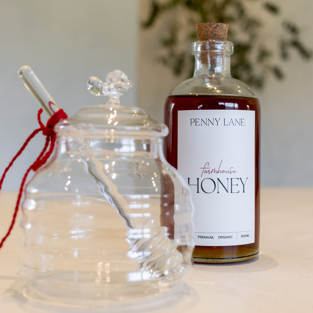 Glass honey jar with glass dipper that has a red tie on it, standing next to a glass bottle of honey with a cork lid in front of greenery