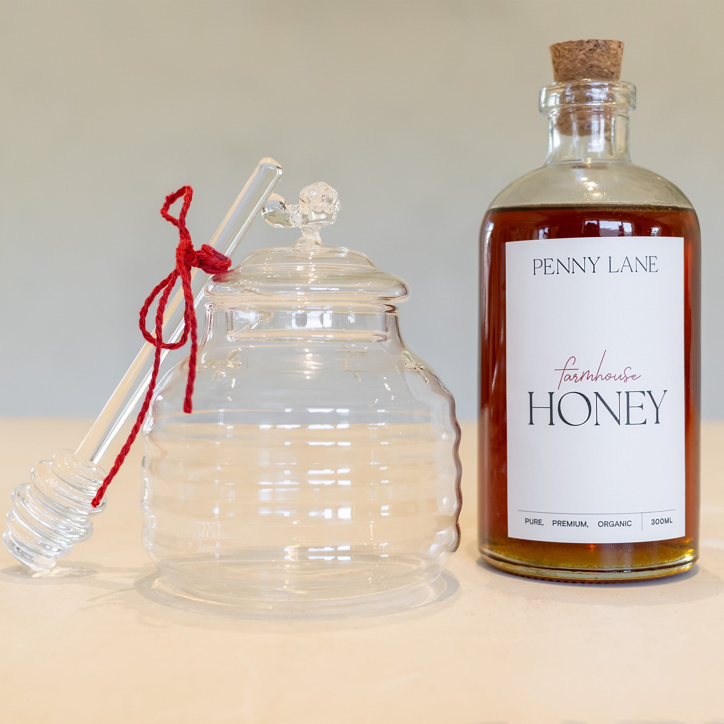 Glass honey jar with glass dipper that has a red tie on it, standing next to a glass bottle of honey with a cork lid
