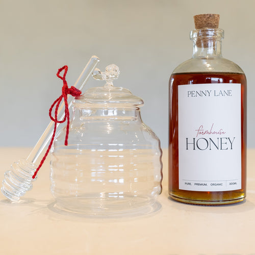 Glass honey jar with glass dipper that has a red tie on it, standing next to a glass bottle of honey with a cork lid
