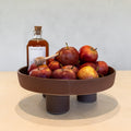 A round brown ceramic pedestal bowl filled with red apples, styled with a bottle of honey and a wooden honey dipper on a light beige tabletop against a neutral gray wall.