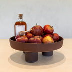 A round brown ceramic pedestal bowl filled with red apples, styled with a bottle of honey and a wooden honey dipper on a light beige tabletop against a neutral gray wall.