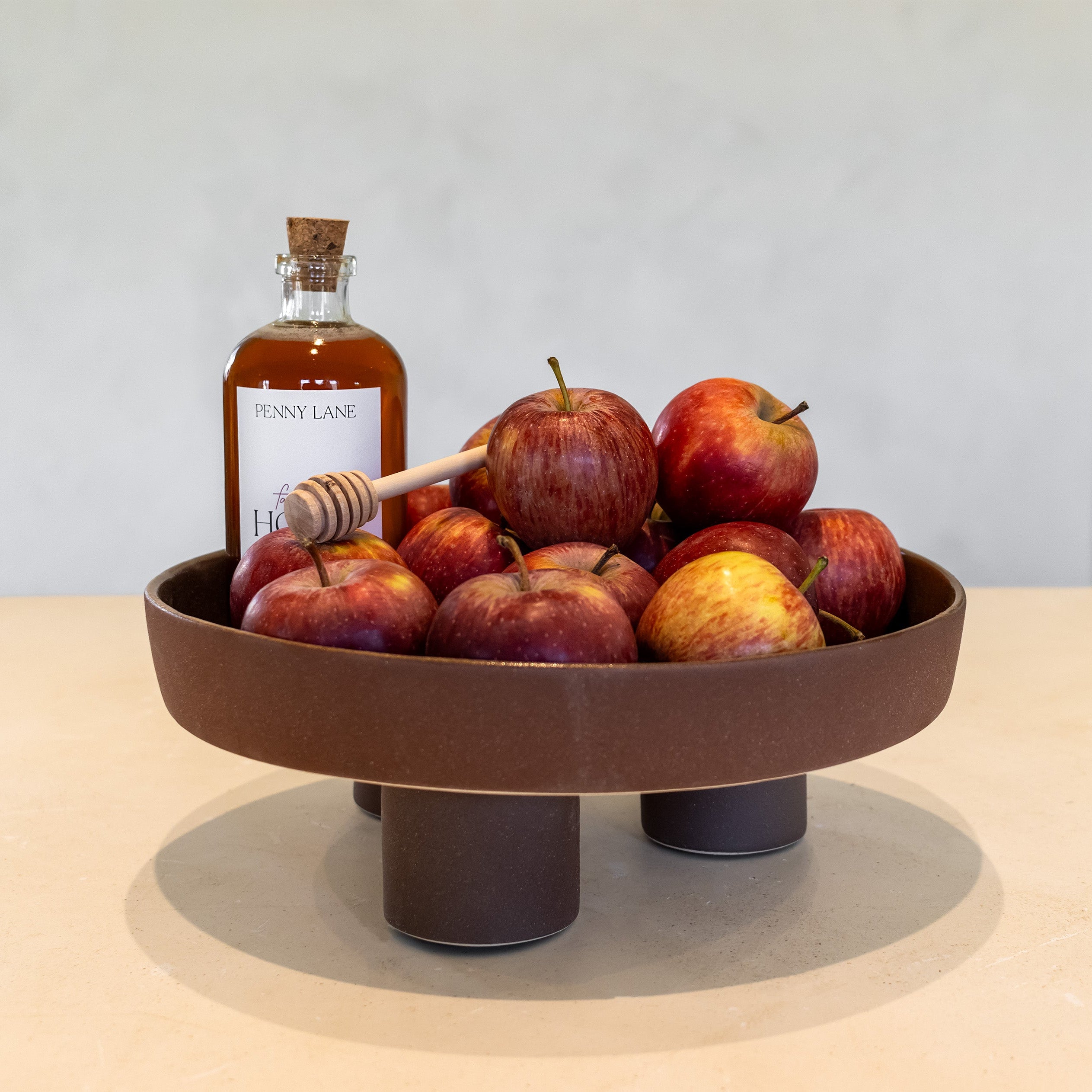 A round brown ceramic pedestal bowl filled with red apples, styled with a bottle of honey and a wooden honey dipper on a light beige tabletop against a neutral gray wall.