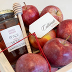 A wooden gift box filled with red apples and a glass jar of Peace of the Home organic honey, tied with a red string and accompanied by a wooden honey dipper and a white tag reading 'Cullinade by Anna'.