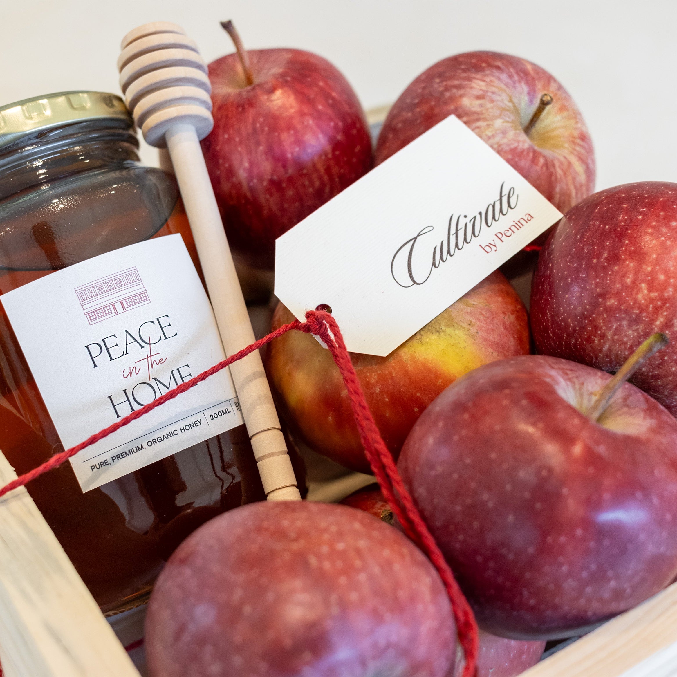 A wooden gift box filled with red apples and a glass jar of Peace of the Home organic honey, tied with a red string and accompanied by a wooden honey dipper and a white tag reading 'Cullinade by Anna'.
