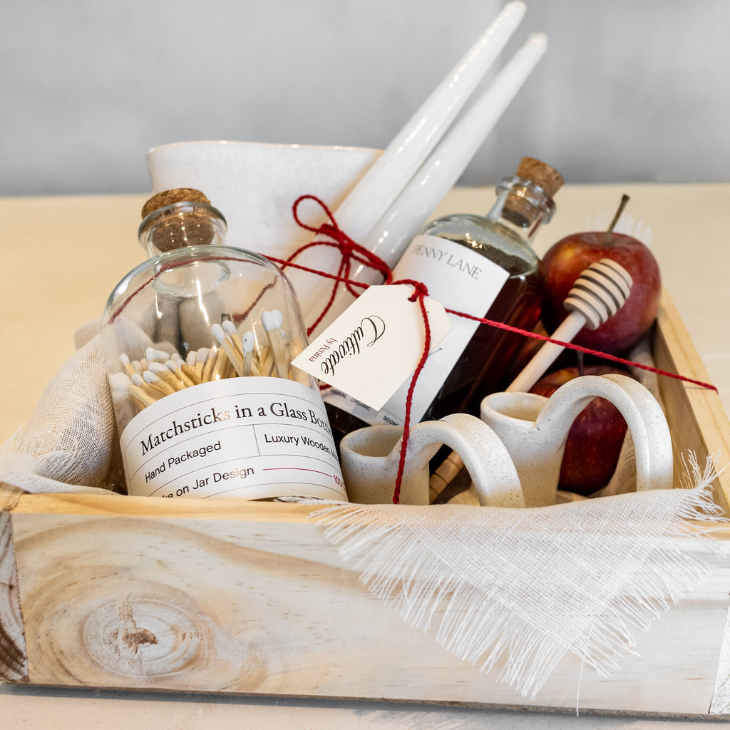 A rustic gift basket arrangement in a whitewashed wooden box, featuring a glass bottle of matchsticks, a small amber bottle, white taper candles, a honey dipper, and a red apple on a light wood surface.