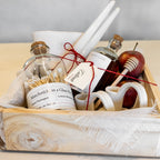 A rustic gift basket arrangement in a whitewashed wooden box, featuring a glass bottle of matchsticks, a small amber bottle, white taper candles, a honey dipper, and a red apple on a light wood surface.