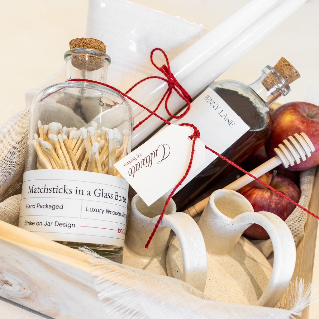 A curated gift box containing a glass bottle of luxury wooden matchsticks, a bottle of Anino Lane balsam, white ceramic cups, a honey dipper, and red apples, arranged in a light wood tray with white fabric.