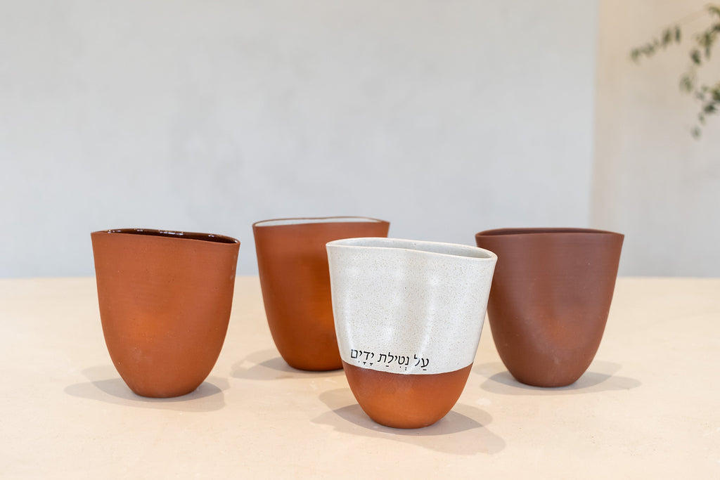 Four ceramic cups arranged on a light tabletop against a soft neutral wall. Three cups are terracotta brown, and the front center cup is white on the upper half with a terracotta brown base and black Hebrew text on the side.