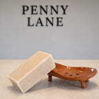A rectangular bar of light beige soap resting on a small brown ceramic soap dish, set against a neutral gray background with the brand name 'PENNY LANE' displayed in black letters on the wall.