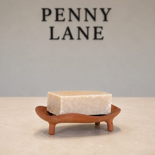 A rectangular bar of soap in a light cream color, displayed on a small curved brown wooden stand against a neutral gray background with the brand name 'PENNY LANE' printed in black at the top.