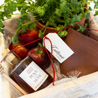 A gift basket filled with fresh green herbs, red apples, a jar of 'Peace Honey' with a wooden dipper, a brown ceramic bowl, and a brown wooden box tied with a red string and a white tag reading 'Candlewood'.