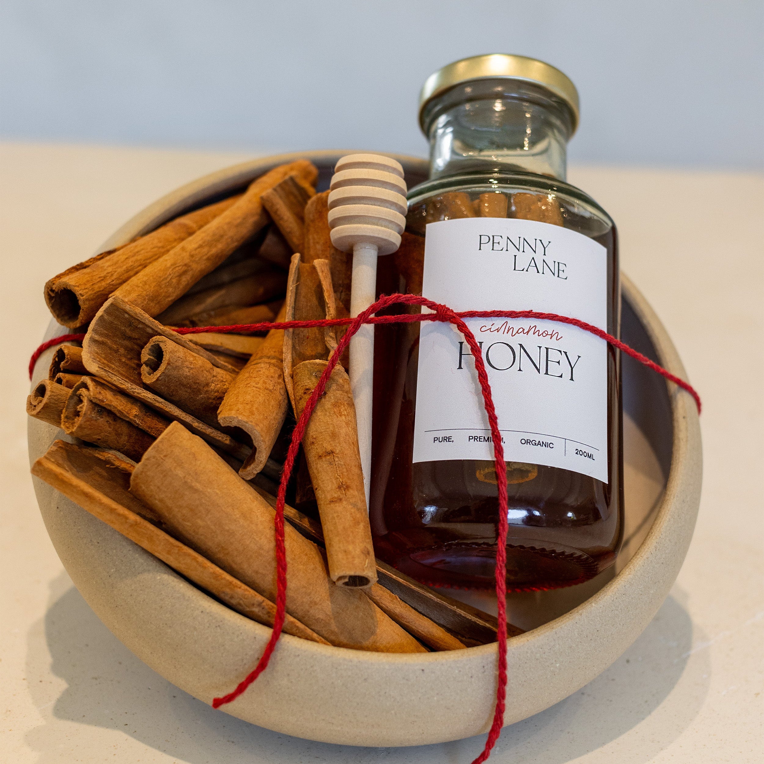 A small glass jar of Penny Lane Honey tied with red string and placed in a ceramic bowl with cinnamon sticks and a wooden honey dipper, on a light surface against a pale blue background.