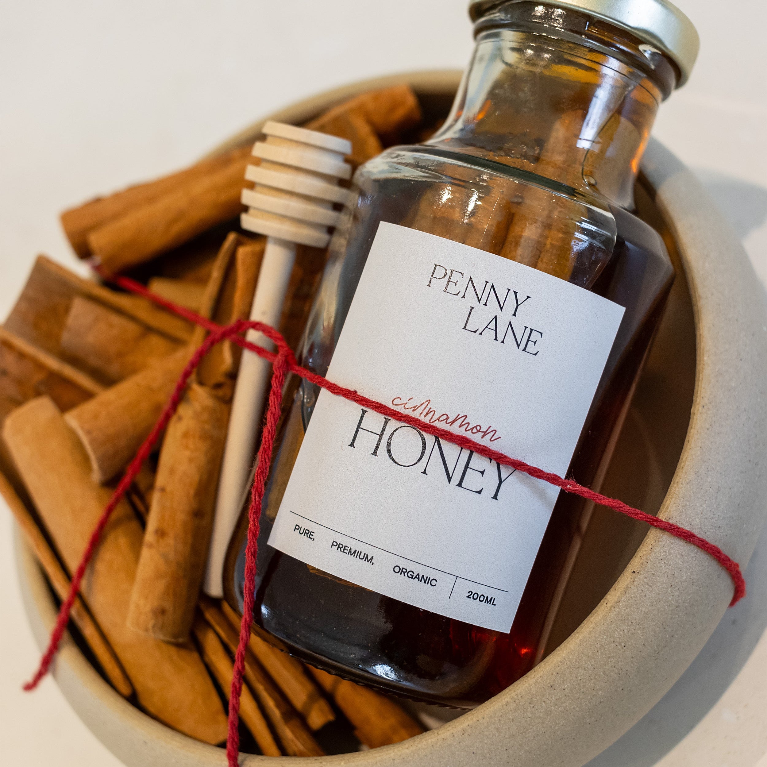 A glass bottle of Penny Lane Citrusation Honey with a gold cap and white label, presented in a ceramic bowl with cinnamon sticks, a wooden honey dipper, and red twine.