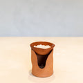 A small terracotta clay candle holder with a wavy cutout design, holding a white candle, set on a light beige surface against a soft blue-gray background.