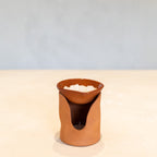 A small terracotta clay candle holder with a wavy cutout design, holding a white candle, set on a light beige surface against a soft blue-gray background.