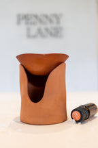 A terracotta clay essential oil burner with a matching bowl on top, shown on a light surface with a small essential oil bottle beside it. The background is a soft off-white with the brand name 'PENNY LANE' blurred at the top.