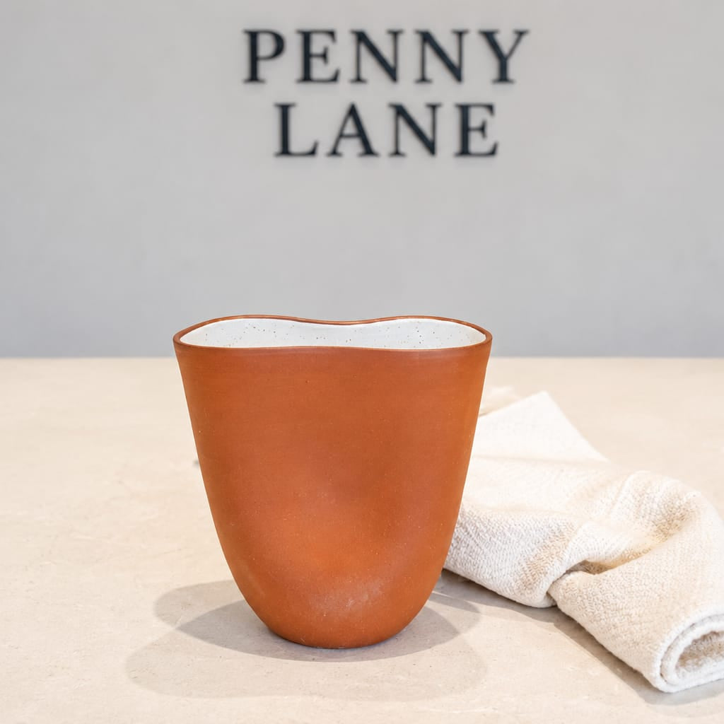 A terracotta-colored ceramic cup with a wavy rim sits on a light beige surface next to a folded white towel, with the brand name 'PENNY LANE' printed on the wall behind it.