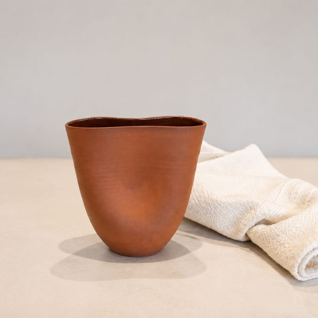 A terracotta-colored ceramic cup with an organic, hand-drawn shape, displayed next to a folded cream-colored towel on a light beige surface against a neutral gray background.