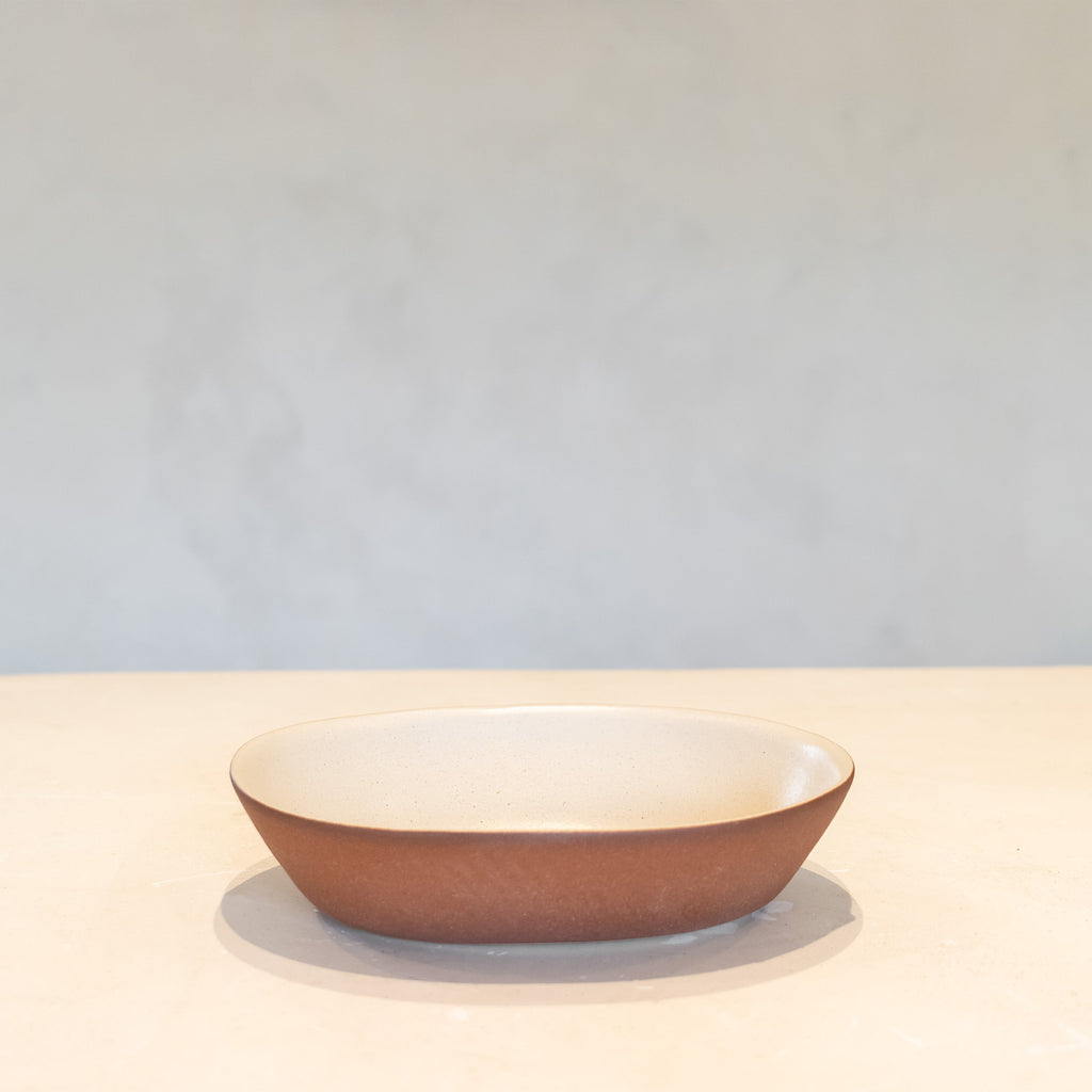 A shallow oval ceramic bowl with a textured terracotta brown exterior and a smooth cream-colored interior, set against a neutral light gray background.