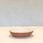 A shallow oval ceramic bowl with a textured terracotta brown exterior and a smooth cream-colored interior, set against a neutral light gray background.