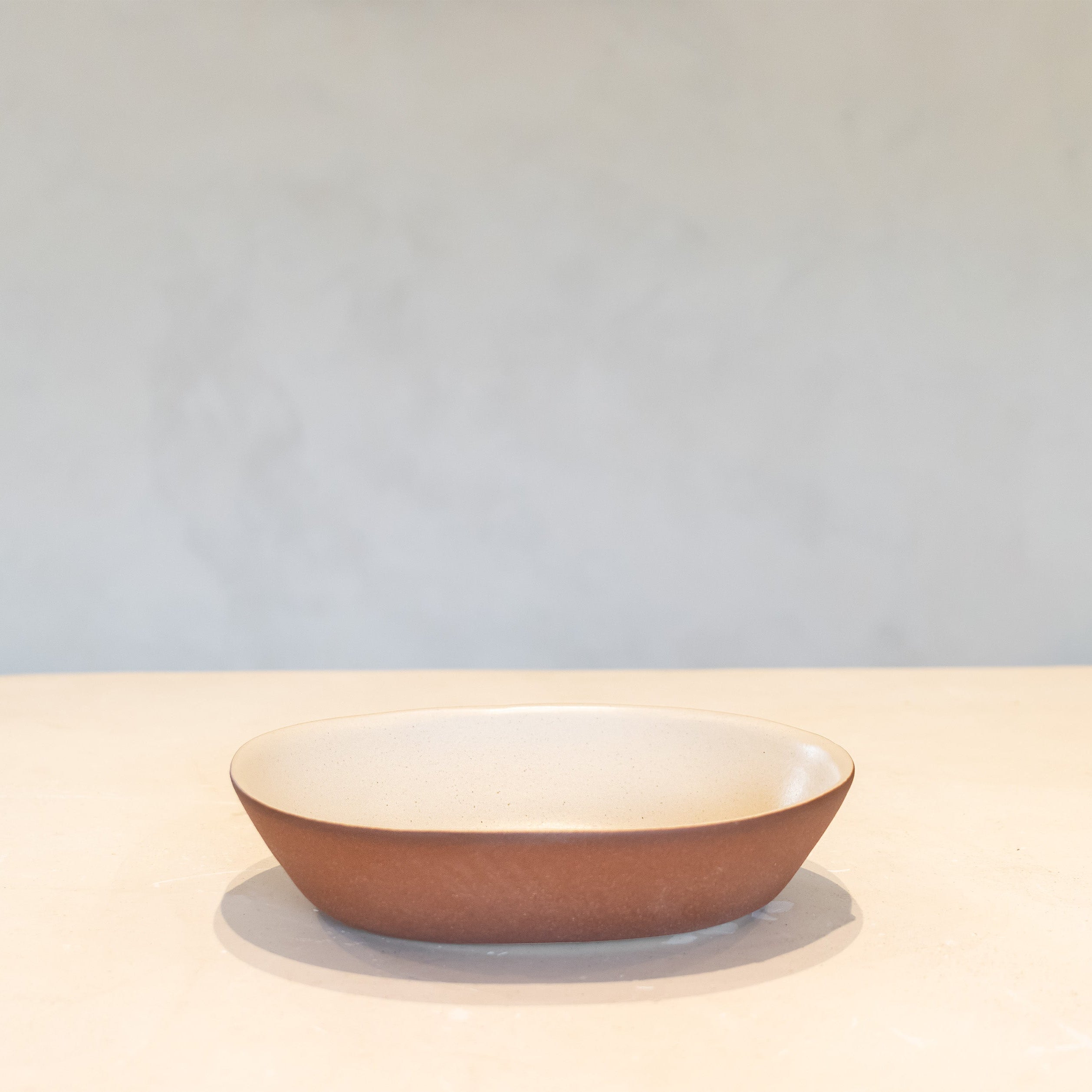 A shallow oval ceramic bowl with a textured terracotta brown exterior and a smooth cream-colored interior, set against a neutral light gray background.