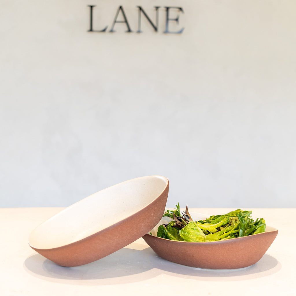 Two shallow oval serving bowls in a matte terracotta finish, one tipped over to show the white interior and one holding a green salad, set on a white surface against a light gray wall with the brand name LANE above.