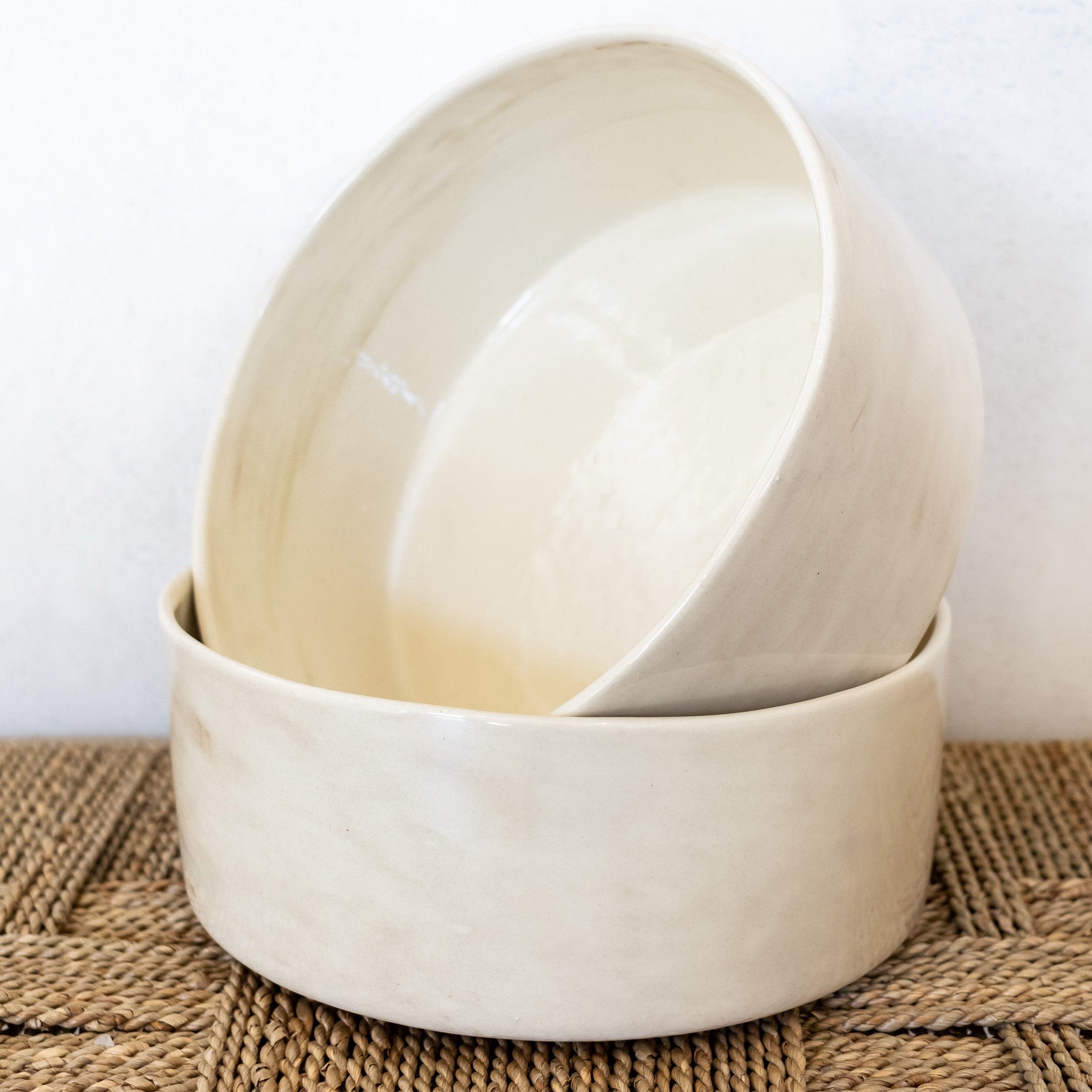 Two beige ceramic bowls stacked on a woven mat with a white background