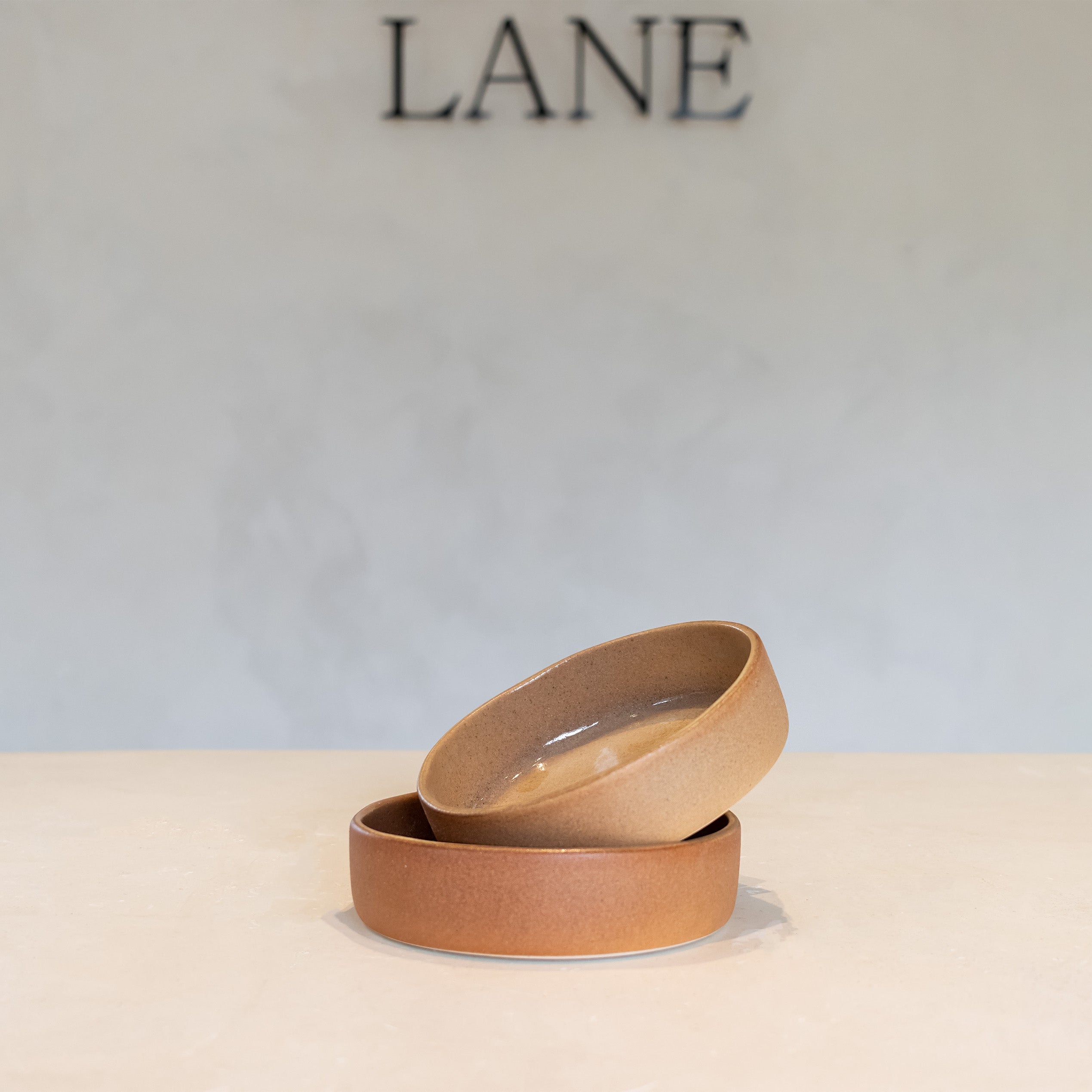 Two small stacked ceramic bowls in a warm tan or light brown glaze, set on a light surface against a pale gray wall with the word LANE visible above.