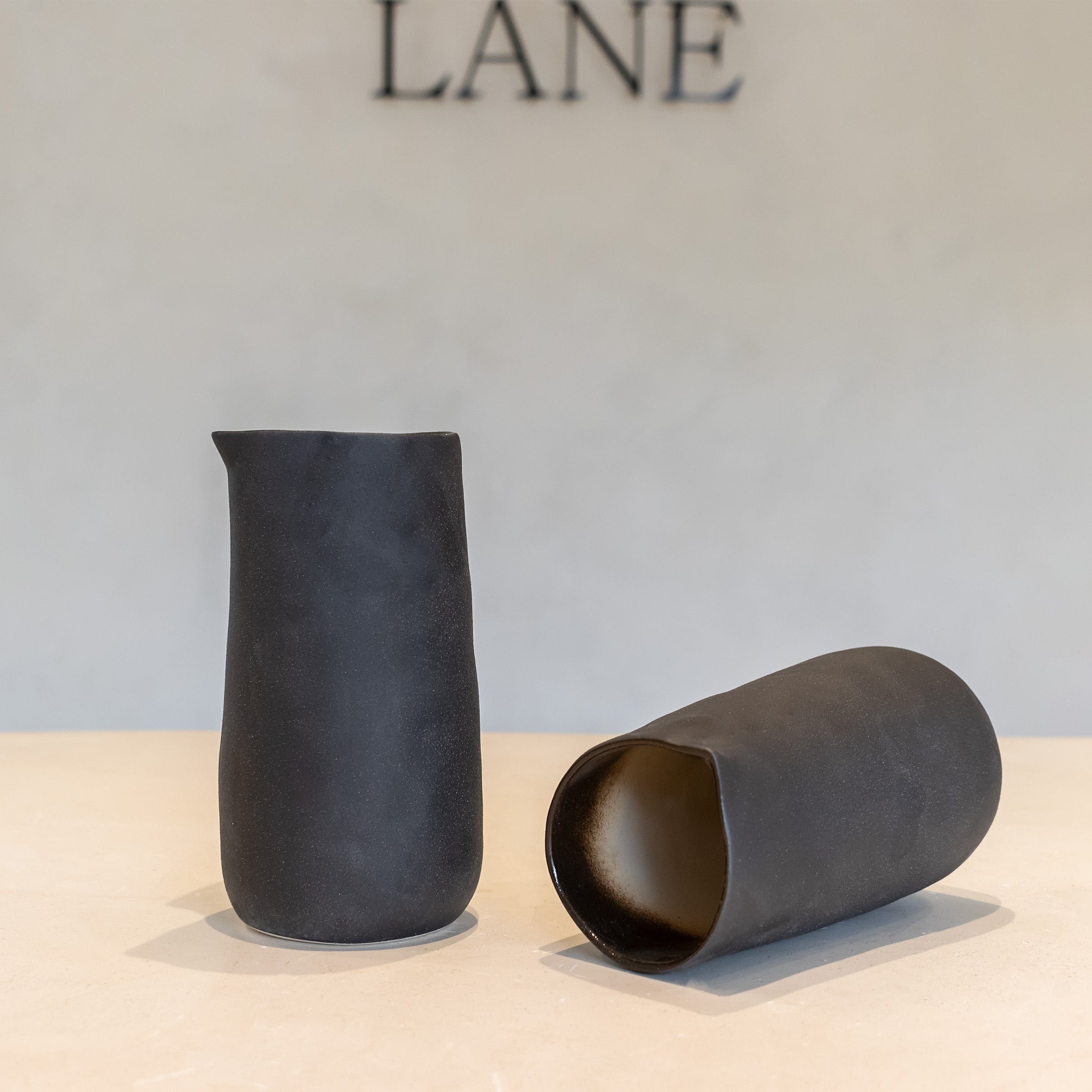 Two matte black ceramic vessels, one standing and one lying on its side, displayed on a light beige surface against a neutral gray wall with the brand name LANE visible at the top.
