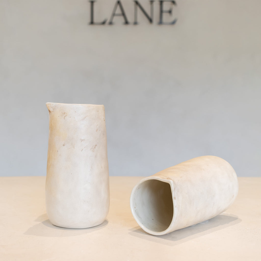 Two small matte ceramic vessels in a light warm off-white tone, one standing upright and one lying on its side, displayed on a tabletop against a soft neutral wall with the word LANE above.