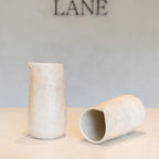 Two small matte ceramic vessels in a light warm off-white tone, one standing upright and one lying on its side, displayed on a tabletop against a soft neutral wall with the word LANE above.