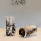 Two cylindrical ceramic vases with a mottled black, white, and gray stone-like glaze, displayed on a light beige surface against a neutral gray background with the brand name LANE partially visible at the top.