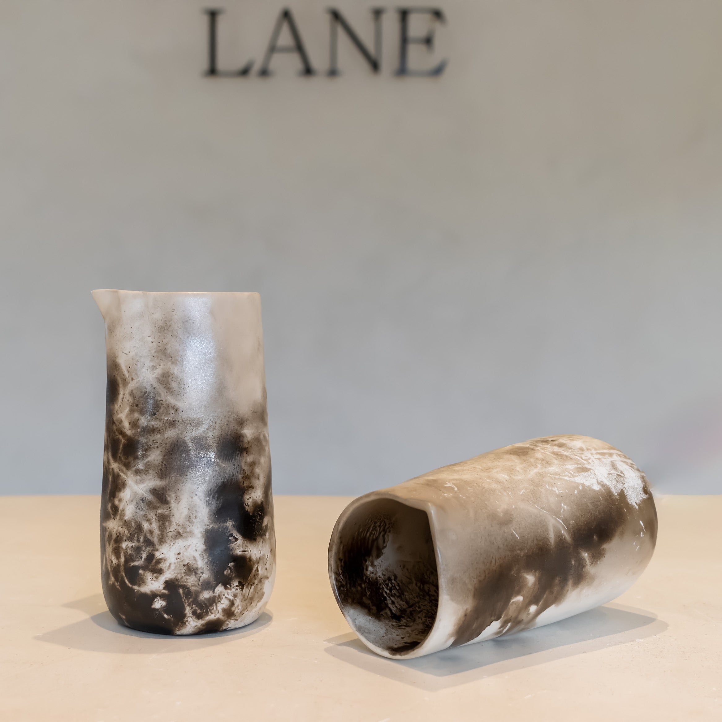 Two cylindrical ceramic vases with a mottled black, white, and gray stone-like glaze, displayed on a light beige surface against a neutral gray background with the brand name LANE partially visible at the top.