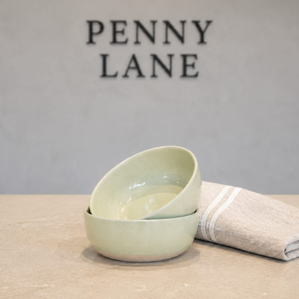 A stack of two pale green ceramic bowls sits on a light stone surface next to a folded beige cloth napkin with white stripes. The background is a textured gray wall with the brand name 'PENNY LANE' in dark lettering.