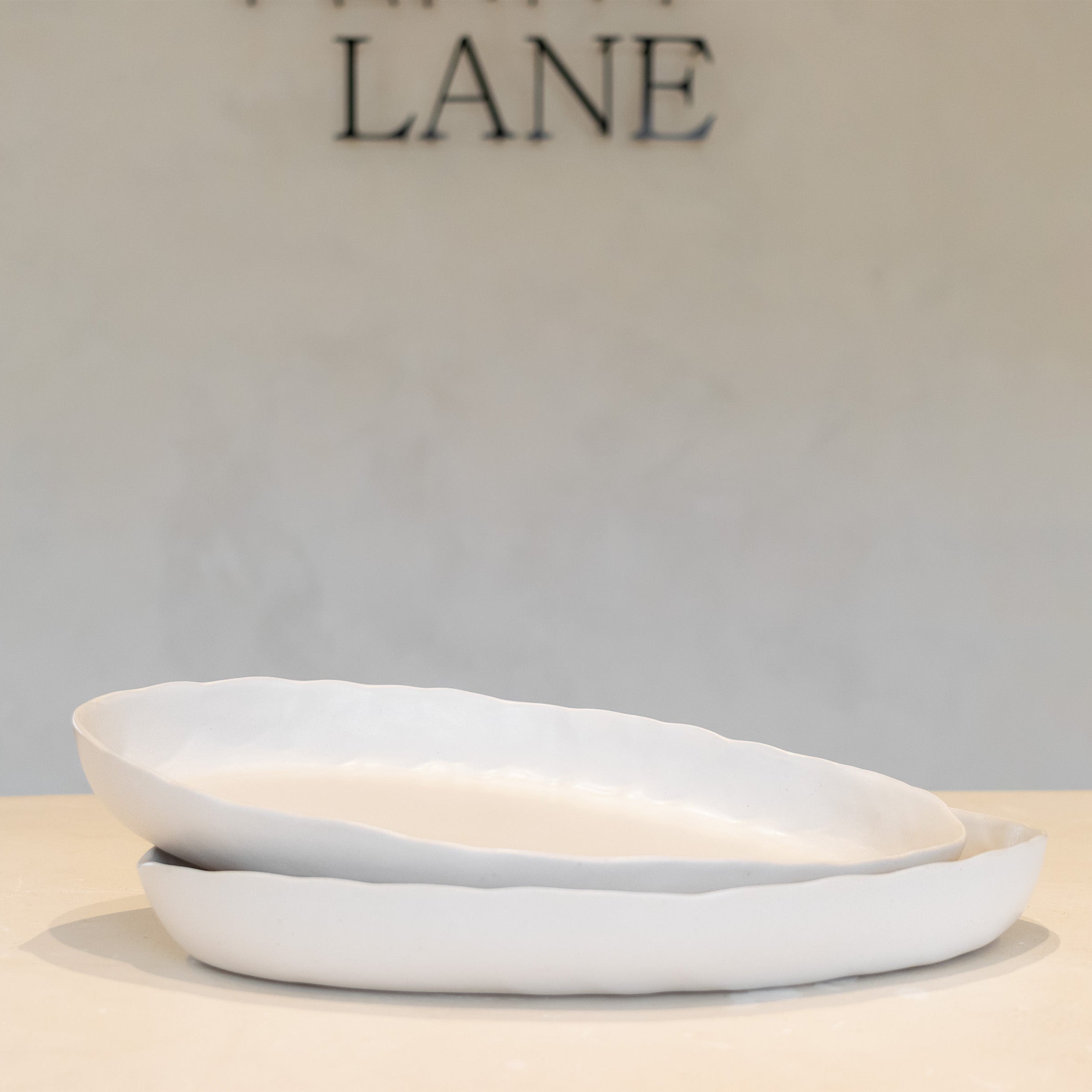 Two stacked white ceramic plates with a scalloped rim, displayed on a light beige surface against a neutral gray wall with 'LANE' partially visible in the background.