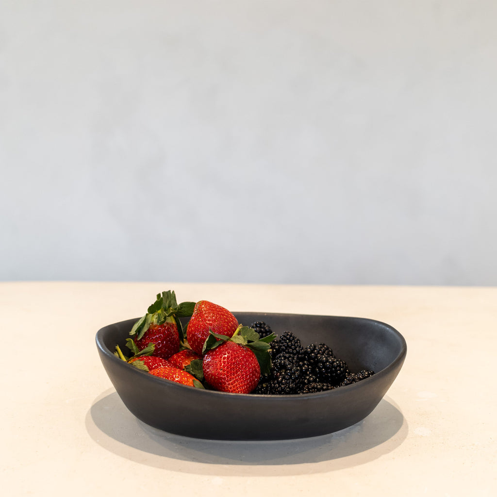 A matte black oval bowl containing fresh red strawberries and blackberries on a light cream surface against a neutral light gray background.