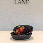 Two stacked matte black ceramic bowls filled with fresh strawberries and blackberries on a light beige surface, with a gray wall and the brand name LANE in the background.