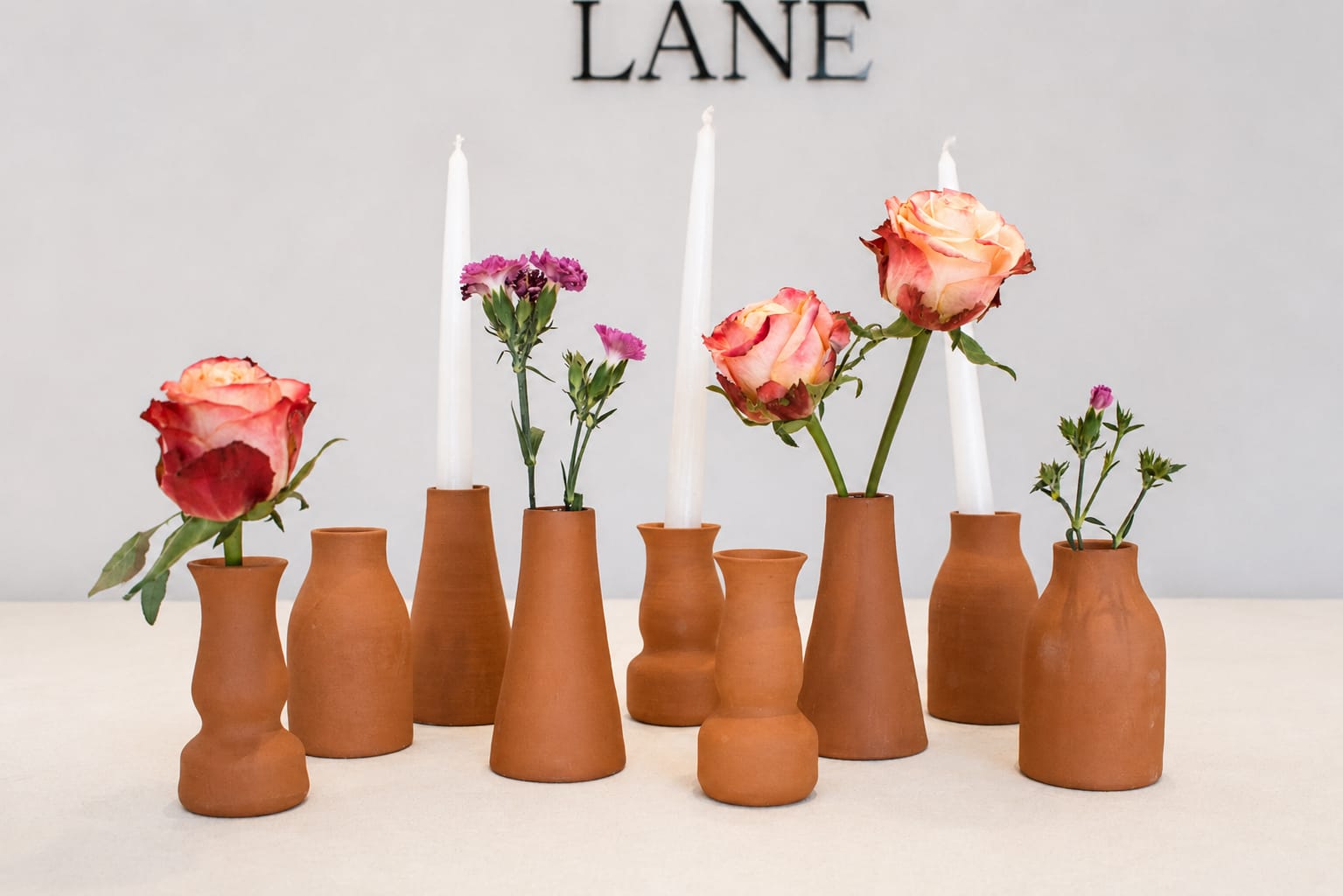 A set of terracotta clay flower vases in various shapes, arranged on a light surface against a light gray wall. Some vases are empty, while others hold white taper candles and small pink and peach roses.