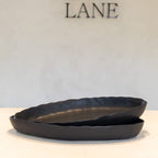 Two overlapping matte black ceramic dishes with a scalloped rim, displayed on a light beige surface against a textured off-white wall with the brand name 'LANE' visible in the background.