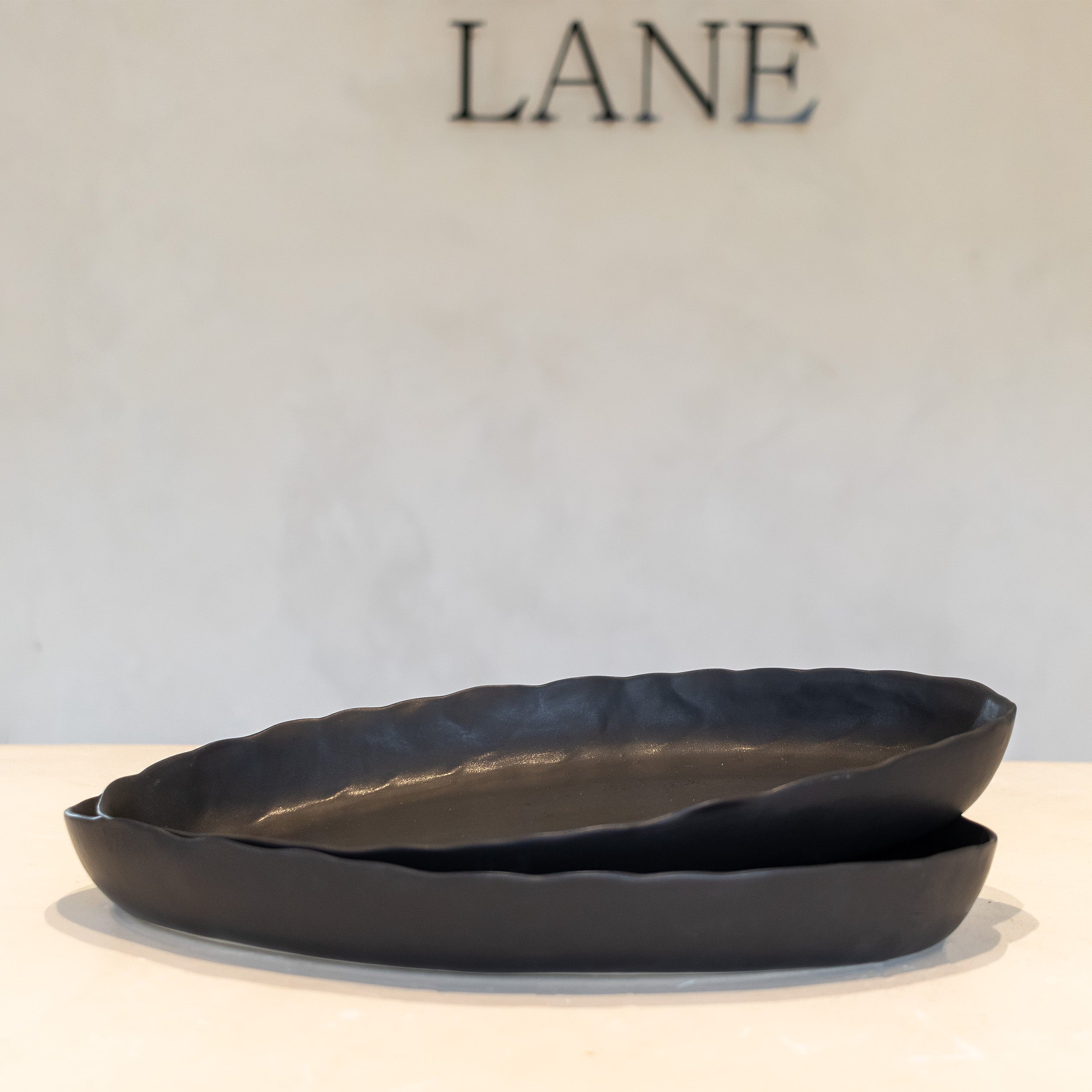 Two overlapping matte black ceramic dishes with a scalloped rim, displayed on a light beige surface against a textured off-white wall with the brand name 'LANE' visible in the background.