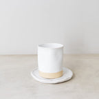 White ceramic cup with beige base on a white plate against a plain background